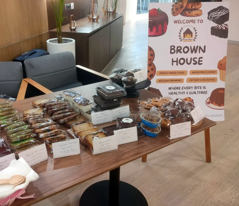 A Brown House pop-up table at an office, with labelled tea cake slices, brownies and cookies beside the Brown House standee